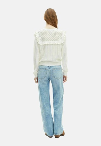Donna con capelli lunghi che indossa un maglione di pizzo bianco con colletto a volant e jeans a vita alta blu chiaro, in piedi di spalle contro uno sfondo bianco.