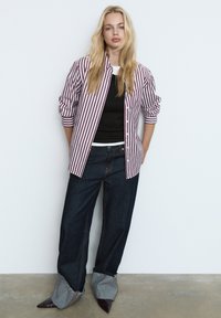 Chemise à rayures rouges et blanches boutonnée sur un haut noir, associée à un jean en denim foncé, avec des poignets effilochés et des chaussures pointues violettes.