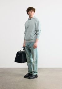 Νεαρός άνδρας που φοράει ανοιχτό γκρι φούτερ με κουκούλα και ξεβαμμένο μπλε τζιν, κρατώντας μια μαύρη δερμάτινη τσάντα και στέκεται σε τσιμεντένιο δάπεδο.