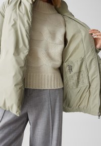Giacca puffer verde chiaro con texture trapuntata, cerniera frontale e etichetta interna. Abbinata a un maglione crema a trecce e pantaloni grigi.