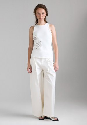 Donna che indossa un top bianco senza maniche con dettaglio in pizzo, pantaloni bianchi larghi e sandali neri, in piedi su sfondo grigio chiaro.