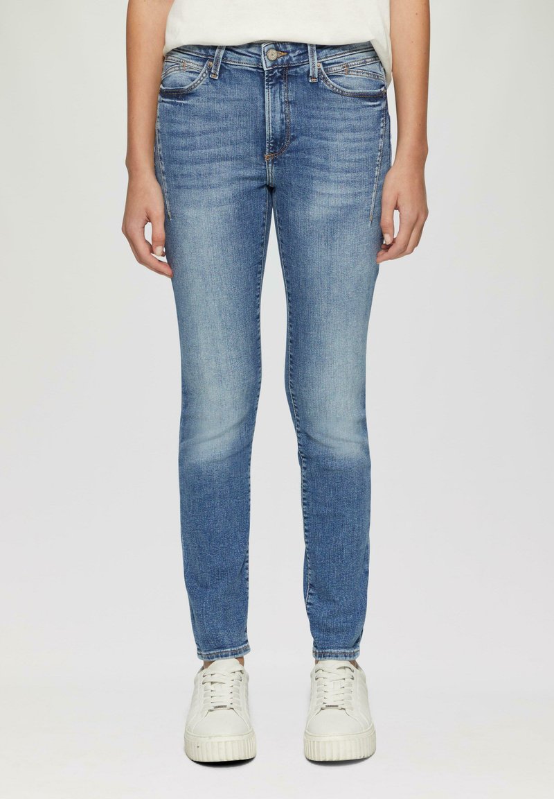 Jeans ajustados en denim azul claro, con una cintura de altura media, diseño de cinco bolsillos y un leve desgaste en las rodillas. Combinados con zapatillas blancas.