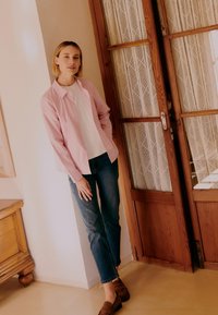 Femeie sprijinită de perete lângă o ușă de lemn cu perdele din dantelă, purtând o cămașă roz cu dungi, un top alb, blugi albastre și mocasini maro.