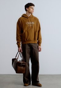 Brun hoodie med vit "CALVIN"-logotyp, framficka, kombinerad med mörkbruna byxor med vida ben och håller en brun läderväska.