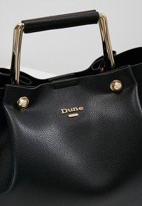 Sort læder håndtaske med en tekstureret overflade, hardware i guldfarve og et topgreb. Har et dekorativt logo på forsiden.