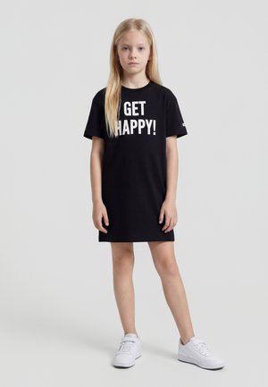 Jeune fille debout portant une robe noire avec l'inscription « GET HAPPY ! » et des baskets blanches sur un fond gris clair uni.