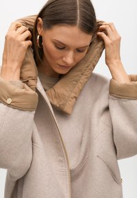 Bež dukserica od flisa s kontrastnim bež pufastim ovratnikom, patentnim zatvaračem i bočnim džepovima. Teksturirana tkanina s glatkim završetkom ovratnika.