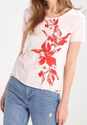 Femme portant un t-shirt rose clair à manches courtes avec un imprimé floral rouge vertical, associé à un jean bleu taille haute.