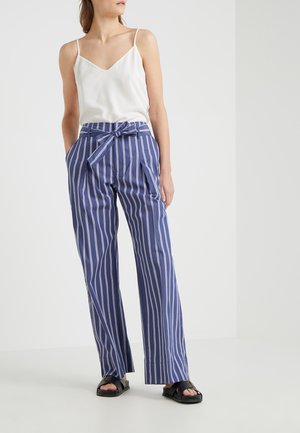 Nainen valkoisessa olkaimettomassa topissa ja korkeavyötäröisissä sinisissä housuissa, joissa valkoiset pystysuorat raidat ja solmittu vyö, seisomassa käsillä taskuissa.