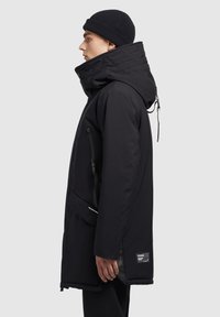 Sort isoleret parka med høj krave, justerbar hætte og dobbelt lynlåslommer. Har en glat tekstur og diskret branding.
