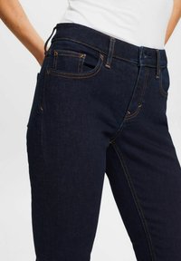 Detailní záběr na osobu oblečenou v tmavě modrých džínech s oranžovým prošíváním a bílém tričku zasunutém do kalhot.