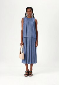 Blaues plissiertes Kleid mit ärmellosem Oberteil und Midilänge, kombiniert mit einer beigen Handtasche und schwarzen Sandalen mit Schnallenakzenten.