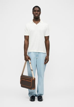 Hombre con polo blanco de manga corta, pantalones vaqueros azul claro, zapatos negros, sosteniendo un bolso cruzado marrón con patrón y una correa beige.