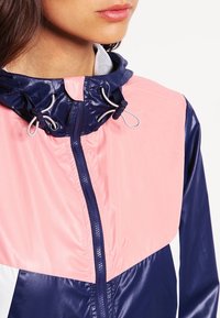 Färgbroderad vindjacka i marinblått, rosa och vitt. Har dragkedja fram, justerbar huva och en slät, glänsande tyg. Silverfärgade detaljer.