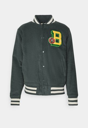 Chaqueta de pana en verde oscuro con puños y dobladillo de canalé a rayas. Presenta una letra "B" bordada y un detalle de parche redondo. Cierre frontal de botones.