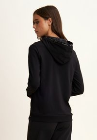 Sudadera negra con cremallera, de textura suave, que cuenta con capucha y detalle del logo en el interior del cuello. Incluye mangas largas y bolsillos laterales.