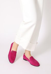 Roze suède instappers met een ronde neus, platte hak en decoratieve metalen accent. Gecombineerd met witte, wijde broek.