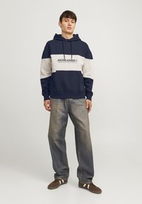 Sudadera bloqueada en color azul marino y gris claro con capucha, que presenta un estampado de logo en la parte frontal. Combinada con vaqueros sueltos y descoloridos y zapatos marrones.