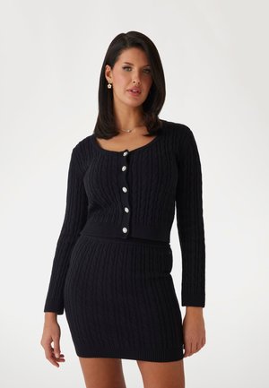 Black knitted cardigan and skirt set featuring cable texture, long sleeves, scoop neckline, and large button accents.