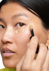 Applicatore di fondotinta in mano, che applica un correttore liquido beige chiaro nella zona sotto gli occhi. Texture della pelle liscia con un finish naturale.