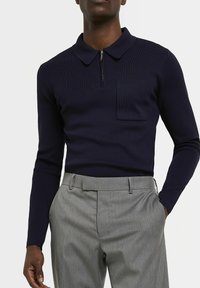 Polo à manches longues bleu marine avec un col zippé, texture côtelée et une poche avant, associé à un pantalon tailleur gris.