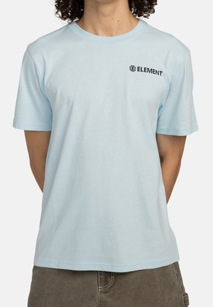 Osoba ubrana w blado niebieską koszulkę z krótkim rękawem z czarnym logo "ELEMENT" na lewej piersi oraz ciemnoszare spodnie, stojąca z rękami za plecami.