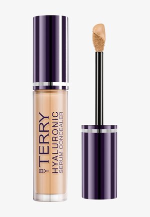 Flacon de correcteur avec bouchon violet foncé et applicateur montrant du correcteur liquide beige au bout.