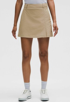 Person iført beige omslagsnederdel, hvide sokker og hvide snøresko stående mod en ensfarvet baggrund.
