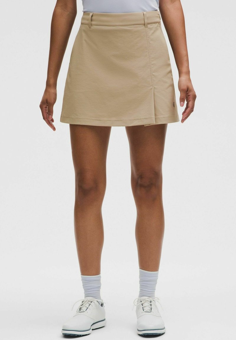 Personne portant une jupe beige portefeuille, des chaussettes blanches et des chaussures blanches à lacets, debout devant un fond uni.