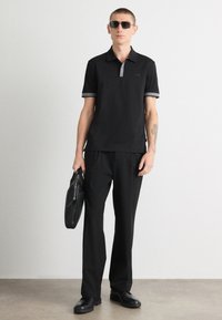 Camisa polo preta com detalhes cinzentos, combinada com calças e sapatos pretos. Modelo segura uma pasta preta e usa óculos de sol escuros. Textura suave.