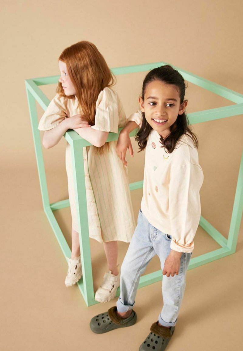 Zwei Kinder stehen neben einem mintgrünen Würfel. Eines trägt ein gestreiftes cremefarbenes Kleid, das andere ein cremefarbenes Oberteil mit bunten Applikationen und blaue Jeans.