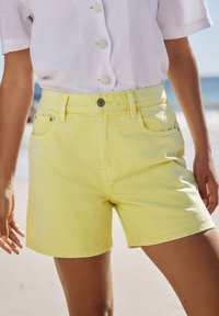 Shorts di denim giallo con vita alta, tasche frontali e chiusura a bottone metallico. Tessuto testurizzato e cuciture visibili.