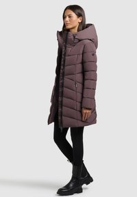 Dolga, podložena bunda v potlačenem vijoličnem odtenku. Ima visok ovratnik, diagonalno prešito zasnovo, sprednji zadrgo in nastavljivo kapuco. Nosa se z črnimi leginami in čevlji.