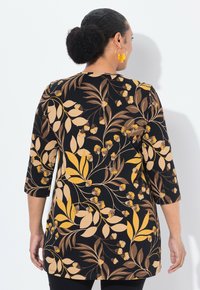 Vrouw met donker haar in een knot, gekleed in een zwarte tuniek met een geel en bruin bloemenpatroon en gele bloemenvormige oorbellen, gezien van achteren.