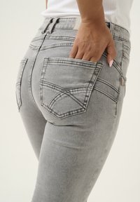 Szürke farmernadrág, testhezálló fazonnal, két hátsó zsebbel és diszkrét varrási részletekkel, oldalnézetből bemutatva.