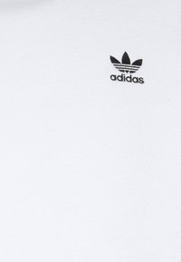 Vit bomull t-shirt med en mjuk textur. Har en svart Adidas-logotyp och ett trebladsmotiv på övre vänstra sidan.