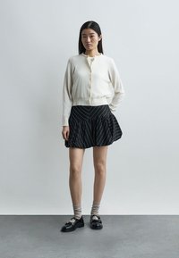 Γυναίκα που στέκεται φορώντας λευκή ζακέτα με κουμπιά, μαύρη φούστα καμπάνα με λευκές ρίγες, μπεζ κάλτσες και μαύρα μοκασίνια μπροστά σε απλό φόντο.