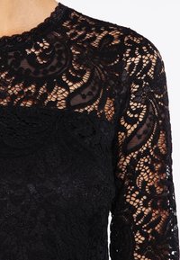 Top de encaje negro con intrincados patrones florales, mangas largas y transparentes, un escote redondo y un panel negro sólido en el pecho.