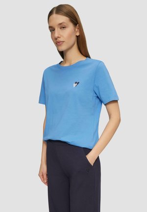 T-shirt en coton bleu clair avec un col rond et des manches courtes. Présente un petit graphic en noir et blanc en forme de cœur sur le côté gauche de la poitrine.