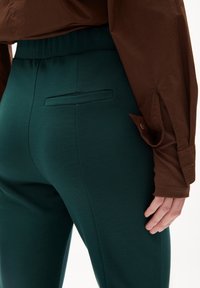 Pantalones verdes ajustados con una textura suave, que cuentan con un bolsillo trasero y una cinturilla elástica. Combinados con una camisa marrón con detalles de botones.