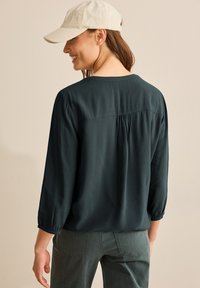 Donkergroene, lichte blouse met 3/4 mouwen, gestructureerde stof en een geplooid detail aan de achterkant. Gecombineerd met grijze broeken en een lichte pet.