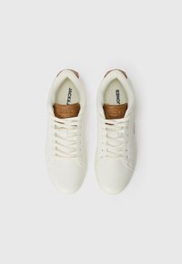 Zapatilla blanca con una parte superior sintética suave, cordones en color crema y acentos de cuero marrón en la lengüeta y el talón. Suela plana de goma.