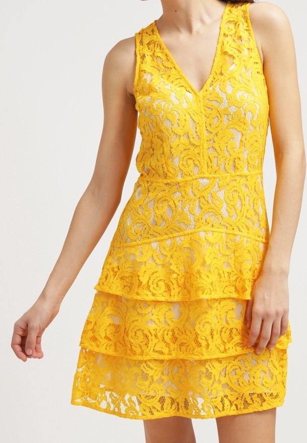 Robe jaune sans manches en dentelle avec col en V et jupe à volants portée par une personne, un bras étendu et l'autre détendu.