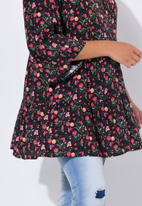 Blouse noire à motif floral avec des fleurs roses et orange, manches cloche et ourlet ondulé, associée à un jean déchiré bleu clair.