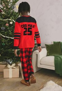 Red and black plaid pajama set featuring long sleeves, bold "COOL YULE 25" text, and coordinating bottoms with checked pattern. Fuzzy slippers complete the look.