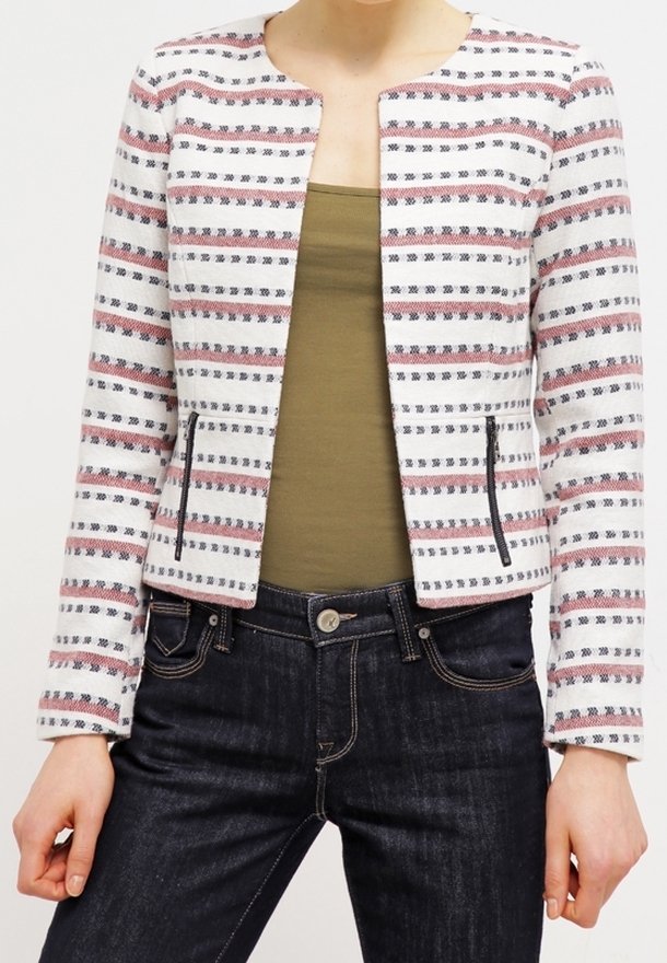 Femme portant une veste blanche à rayures ouverte avec des poches zippées, un haut vert olive et un jean bleu foncé, debout les mains détendues.