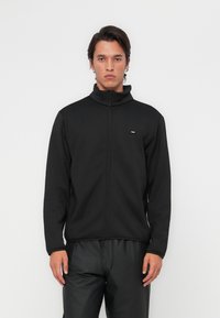 Black zip-up jacket with high collar, made of smooth fabric. Features a small logo on the chest and long sleeves. Worn with black pants.