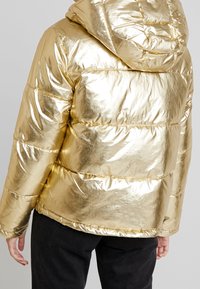 Gouden pufferjack met een glanzend, reflecterend oppervlak, voorzien van een capuchon, doorgestikt ontwerp en elastische manchetten, gedragen over een donkere broek.