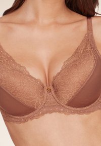 Bra in braun mit floralen Mustern, einem gerippten Stoffabschnitt und einem kleinen dekorativen Knopf in der Mitte des Brückenbereichs.