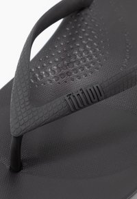 Tongs noires avec un semelle texturée, une lanière lisse avec un motif reptilien et un logo en relief sur la lanière.